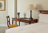 A hotel room with a bed, a desk with a lamp, a chair, a vase with flowers, and a framed picture on the wall.