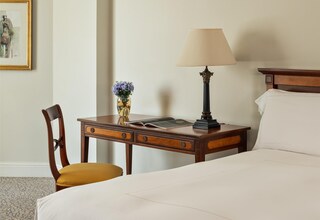 A hotel room with a bed, a desk with a lamp, a chair, a vase with flowers, and a framed picture on the wall.