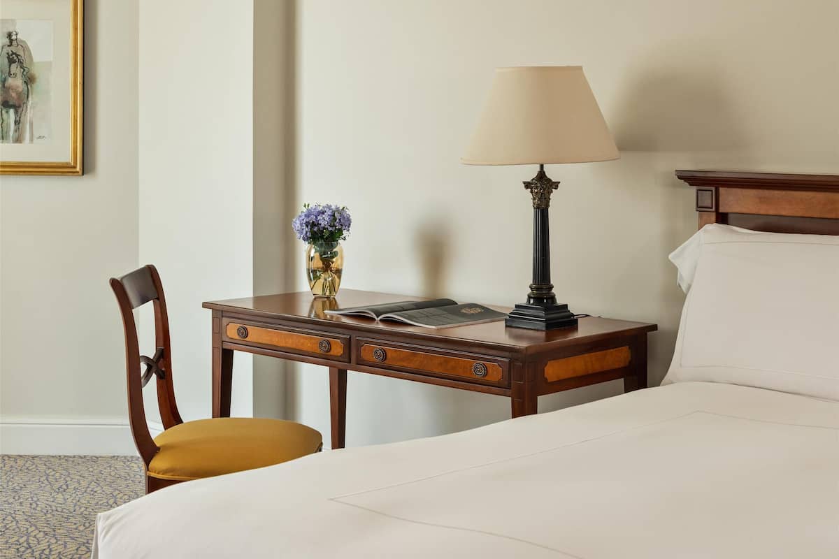 A hotel room with a bed, a desk with a lamp, a chair, a vase with flowers, and a framed picture on the wall.