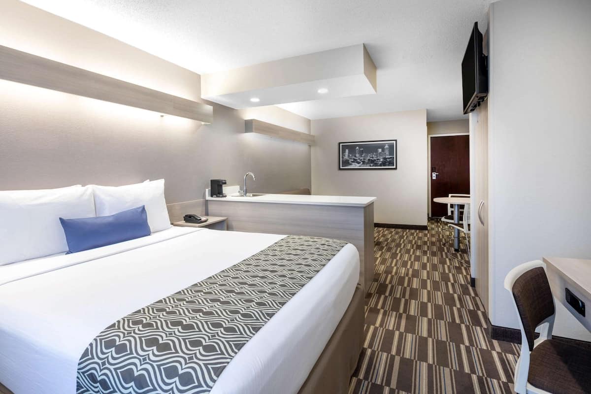 A hotel room with a large bed, a desk, a chair, a small kitchenette, and a TV mounted on the wall.
