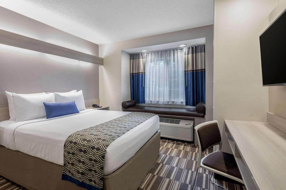 A hotel room with a bed, a desk, a chair, a window with curtains, and a television.