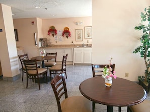 A dining area with a round table and chairs, a kitchen in the background, and a potted plant. - Americas Best Value Inn And Suites Mcdonough (McDonough)