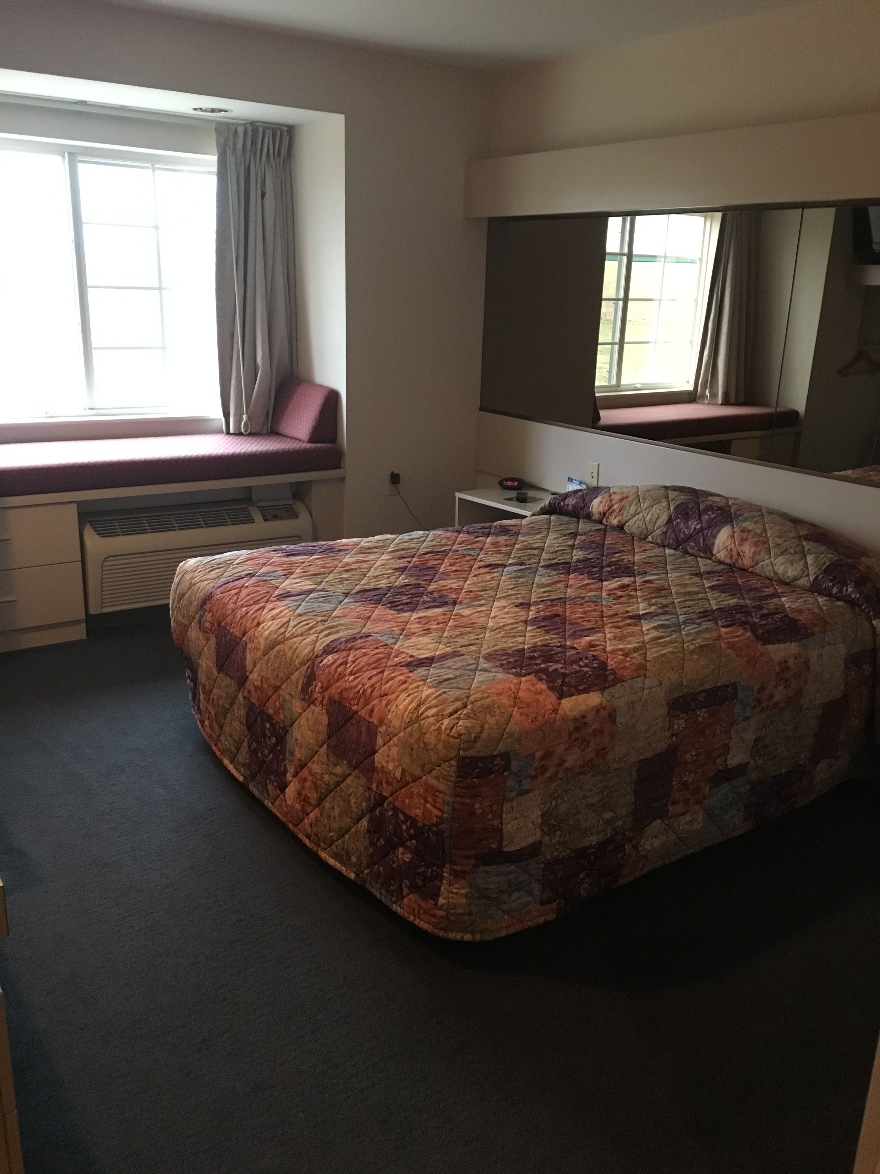 A bedroom with a large bed, a window with curtains, a desk, and a mirror.