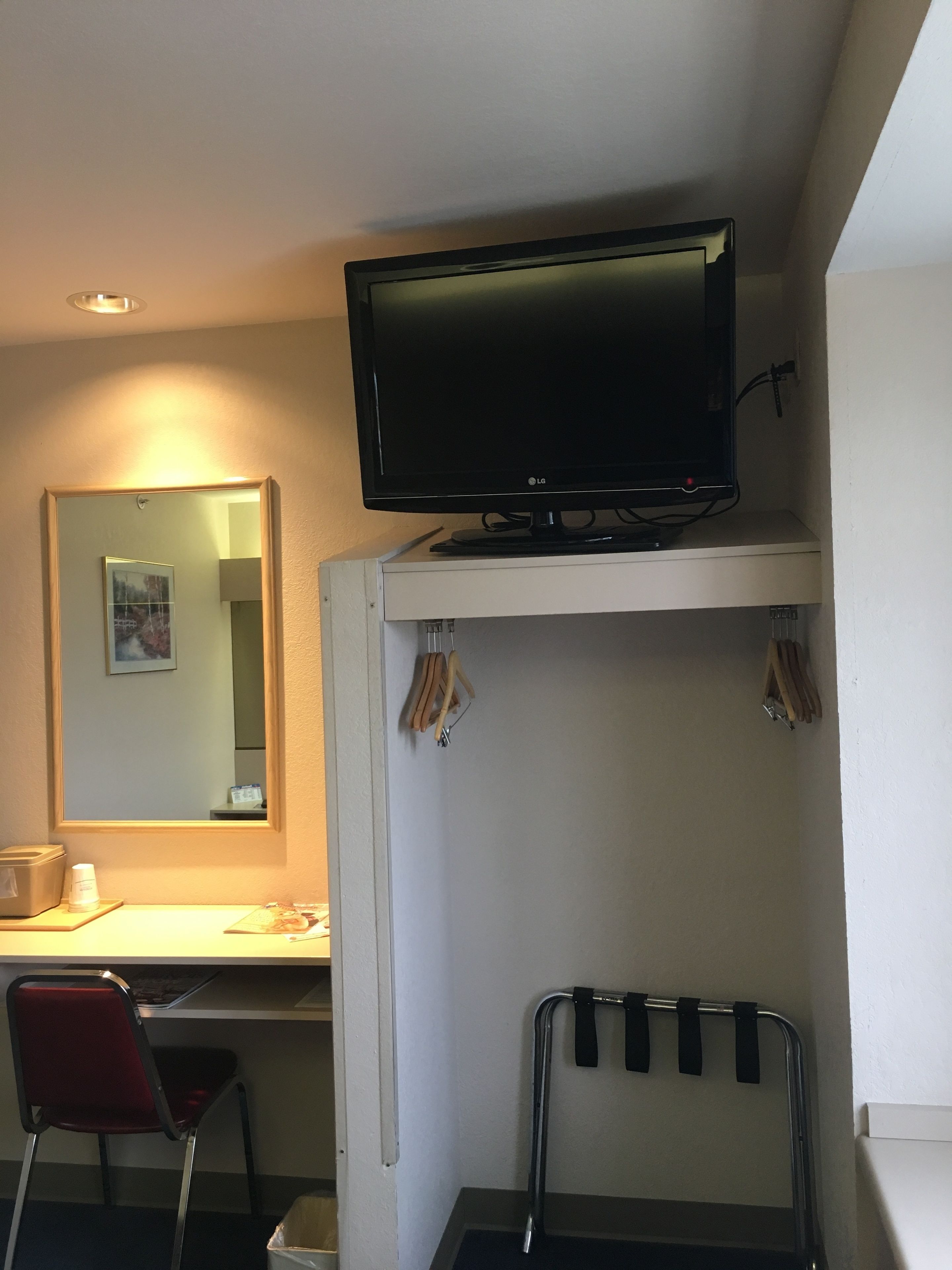 Una habitación pequeña con un escritorio, una silla, un televisor montado en la pared, un espejo y un perchero.