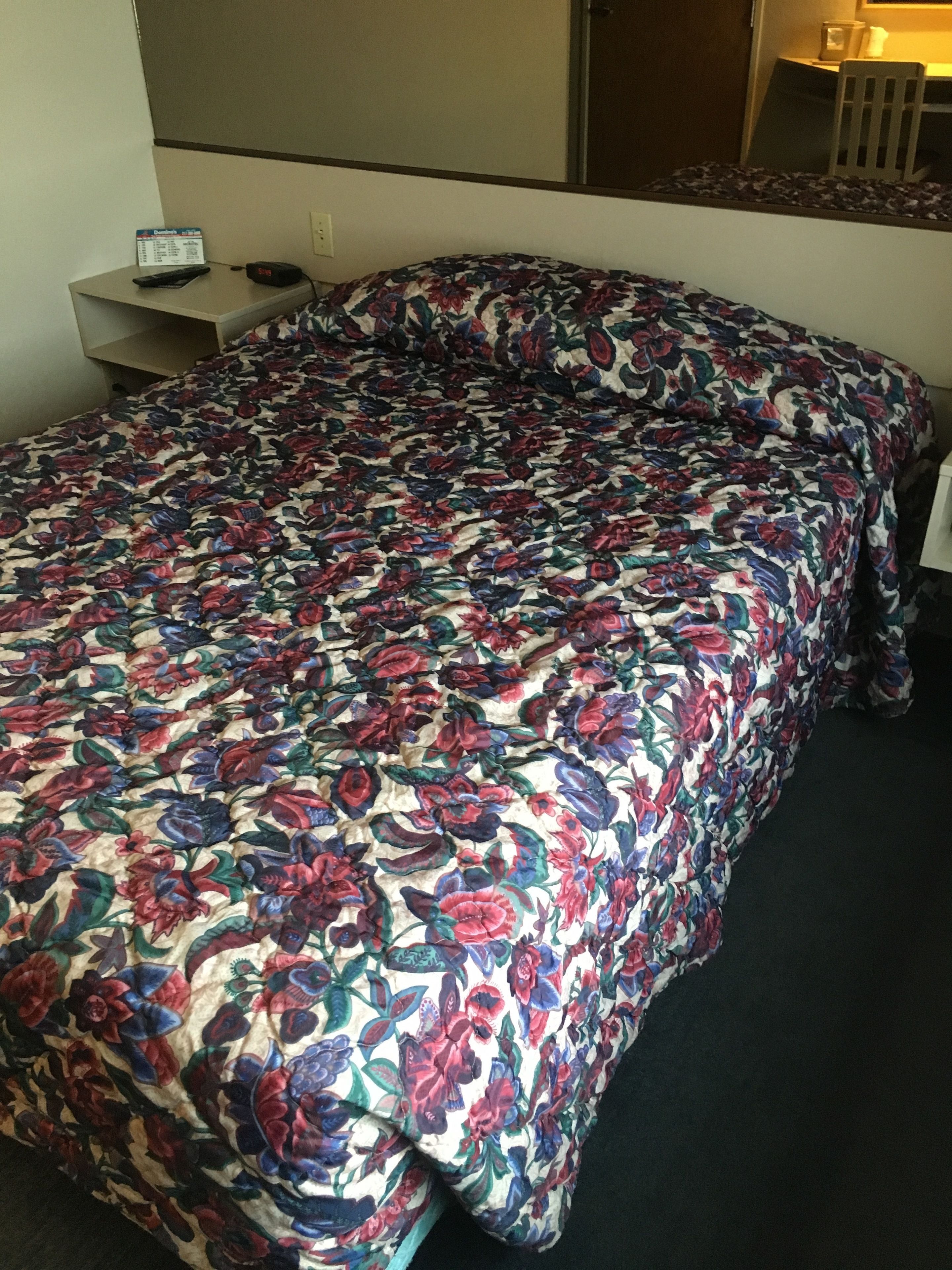 A bed with a floral bedspread, a bedside table with a clock, and a chair in a hotel room.