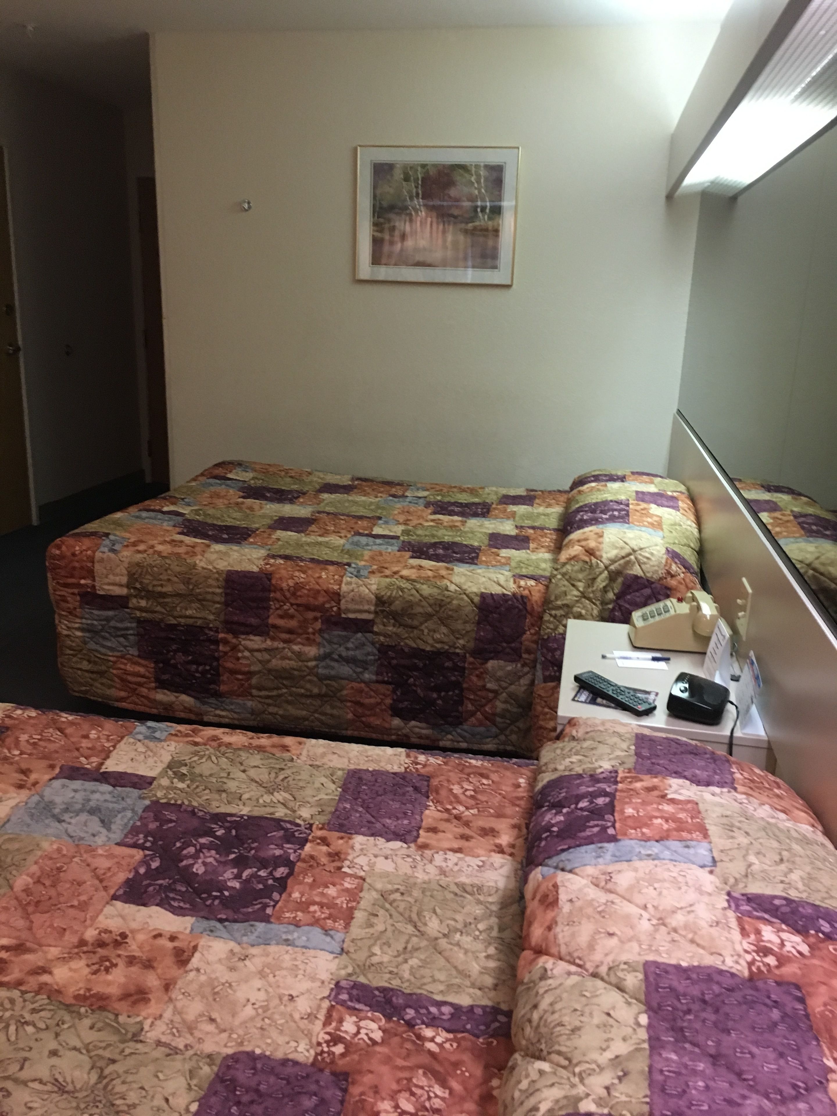 Een hotelkamer met twee bedden, overdekt met kleurrijke quilts, een nachtkastje met een afstandsbediening, een pen en een klein elektronisch apparaat.