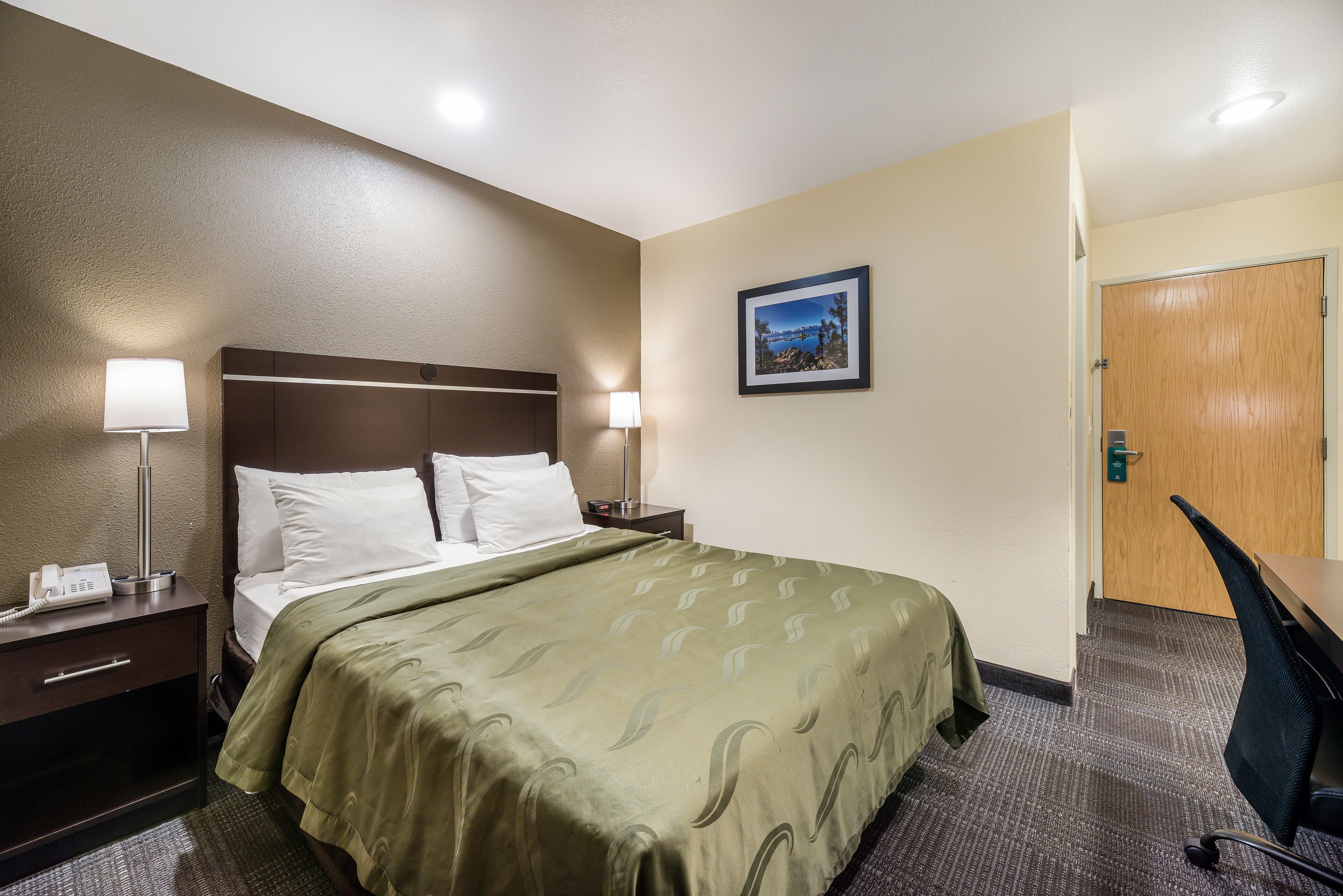 A hotel room with a bed, bedside lamps, a desk, and a door.