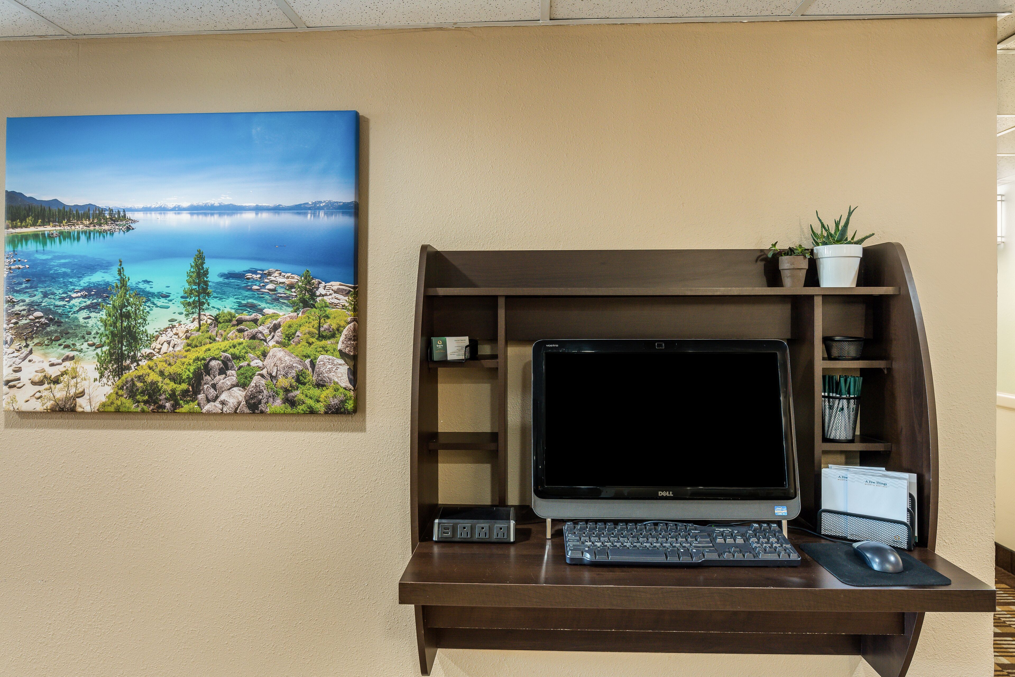 A desk with a laptop, keyboard, and mouse, a potted plant, and a framed landscape painting.