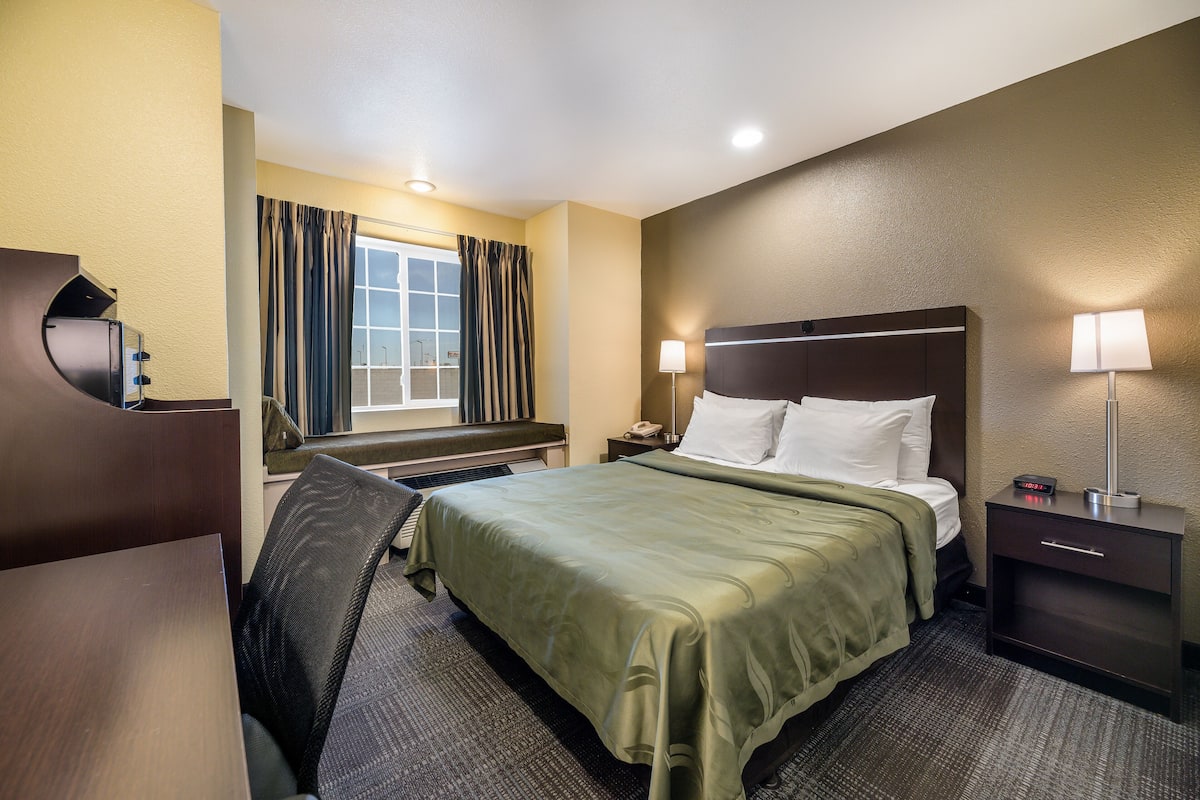 A hotel room with a bed, a desk, a chair, a nightstand, and a lamp.