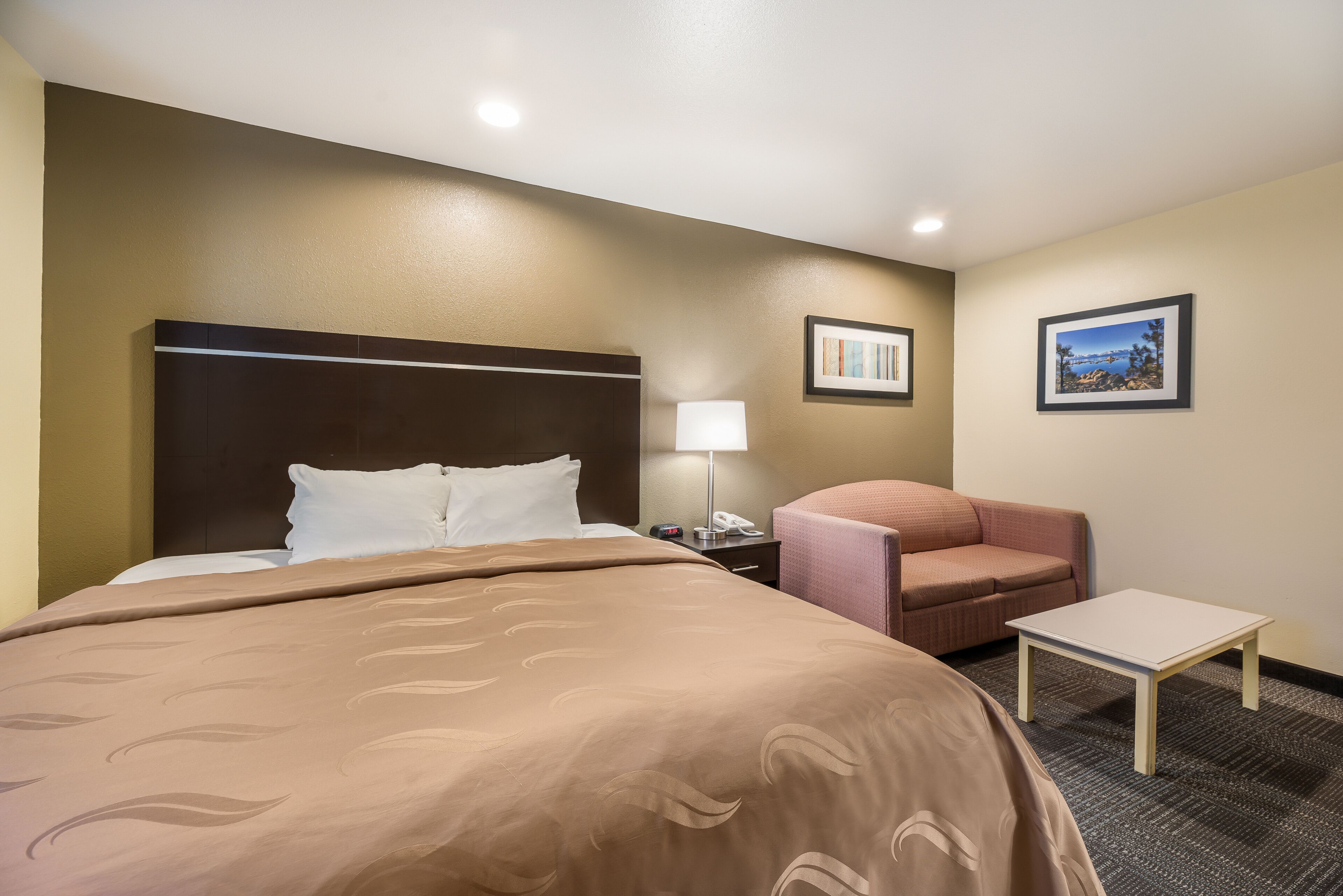 Habitación de hotel con una cama grande, una mesita de noche con lámpara, una pequeña mesa, una silla y dos cuadros en la pared.