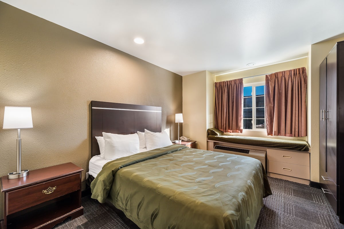 A hotel room with a bed, bedside table, lamp, dresser, and window with curtains.