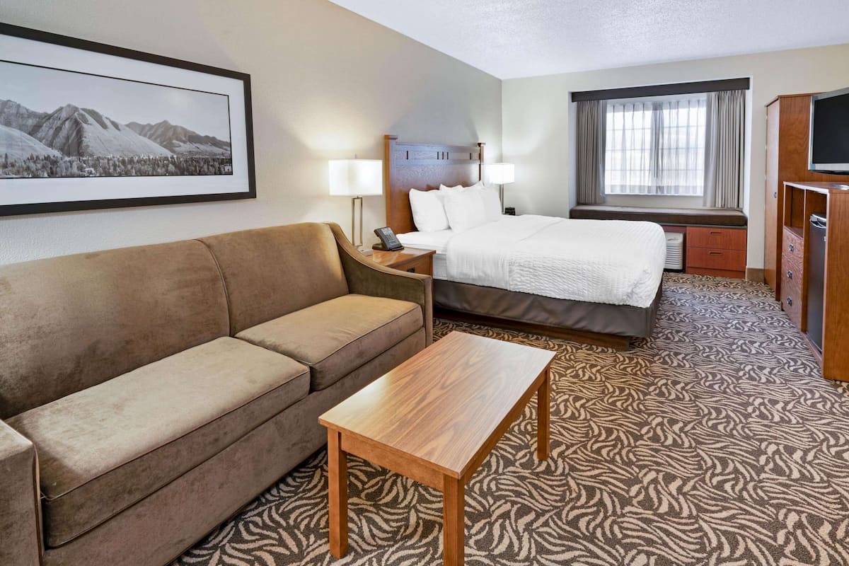 A hotel room with a bed, a sofa, a wooden table, a TV, and a window with curtains.