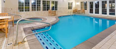 An indoor swimming pool with tiled flooring, a pool ladder, and several chairs around the pool area.