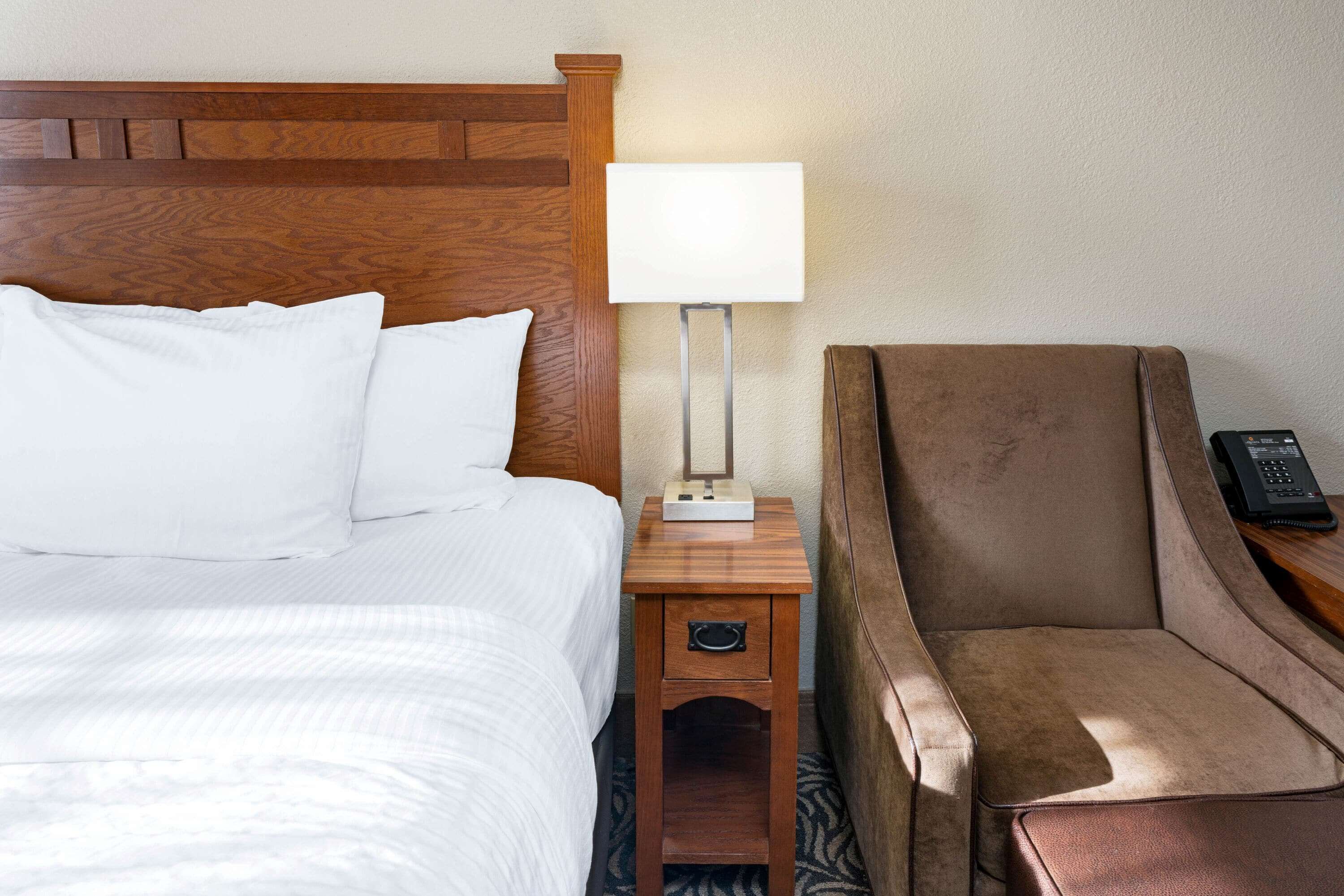 Una habitación de hotel con cabecera de madera, cama con sábanas blancas, mesita de noche con lámpara y un sillón marrón.