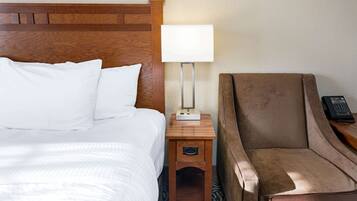 Una habitación de hotel con cabecera de madera, cama con sábanas blancas, mesita de noche con lámpara y un sillón marrón.