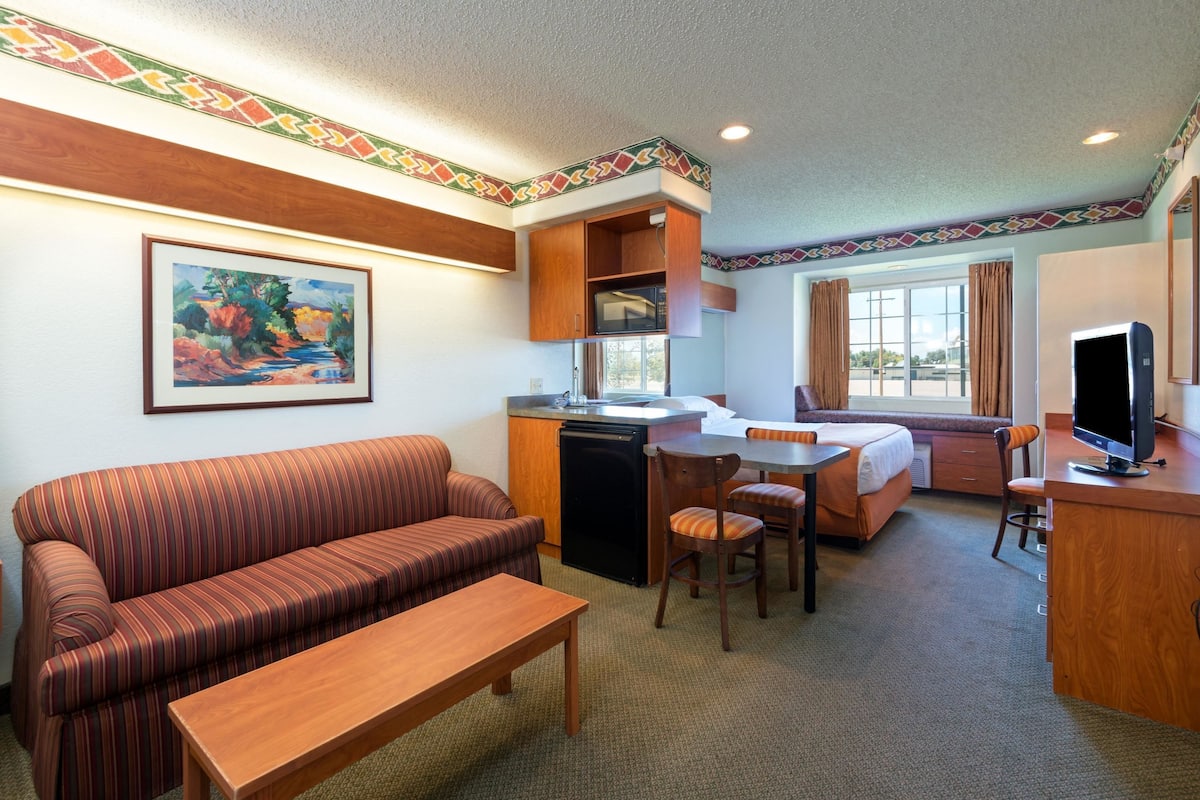 A hotel room with a sofa, dining table, chairs, a small kitchen area, and a television.