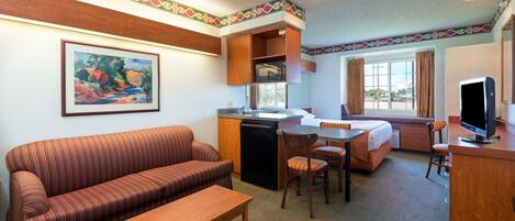 A hotel room with a sofa, dining table, chairs, a small kitchen area, and a television.