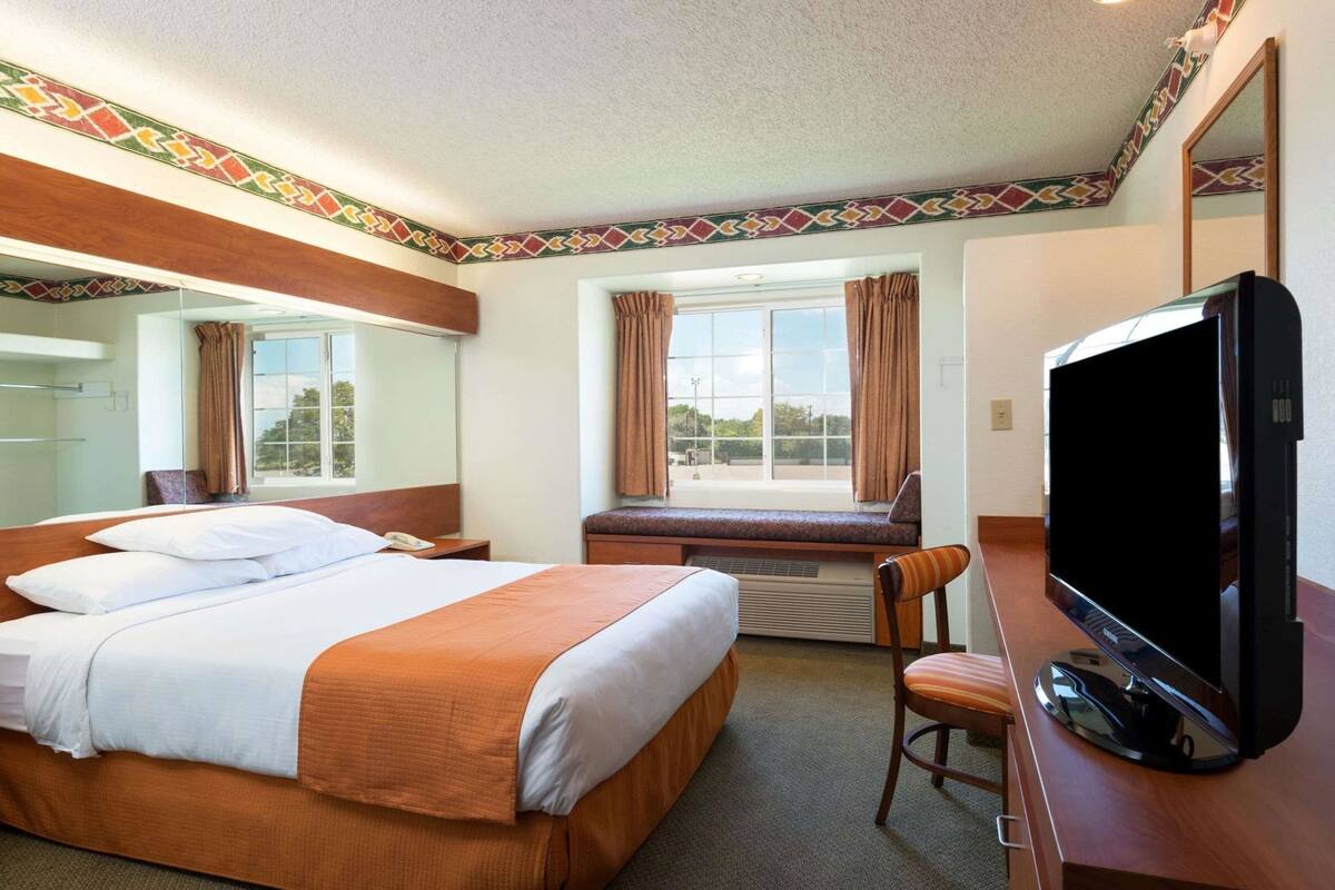 Una habitación de hotel con cama, televisión, ventana con cortinas, un banco y un espejo.