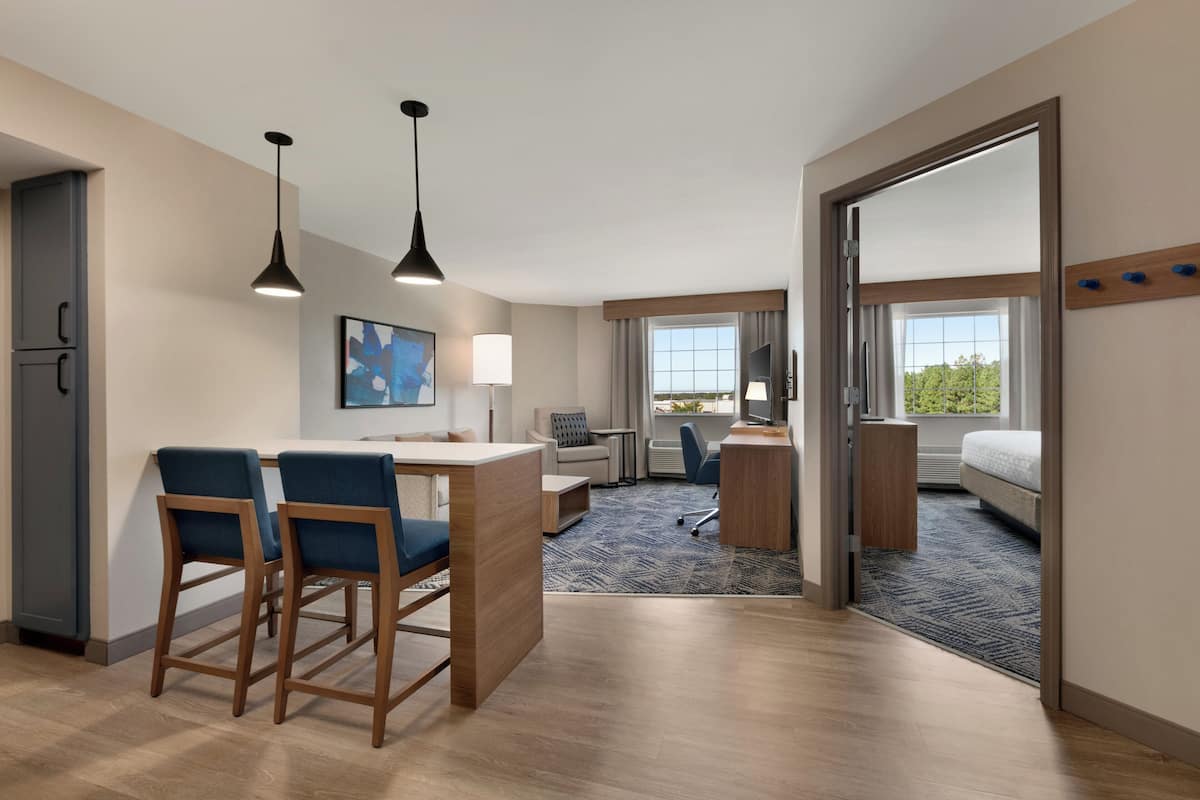 A modern hotel room with a kitchenette, a dining area with bar stools, a large bed, a desk with a chair, and a view of the outdoors.