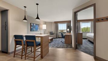 A modern hotel room with a kitchenette, a dining area with bar stools, a large bed, a desk with a chair, and a view of the outdoors.