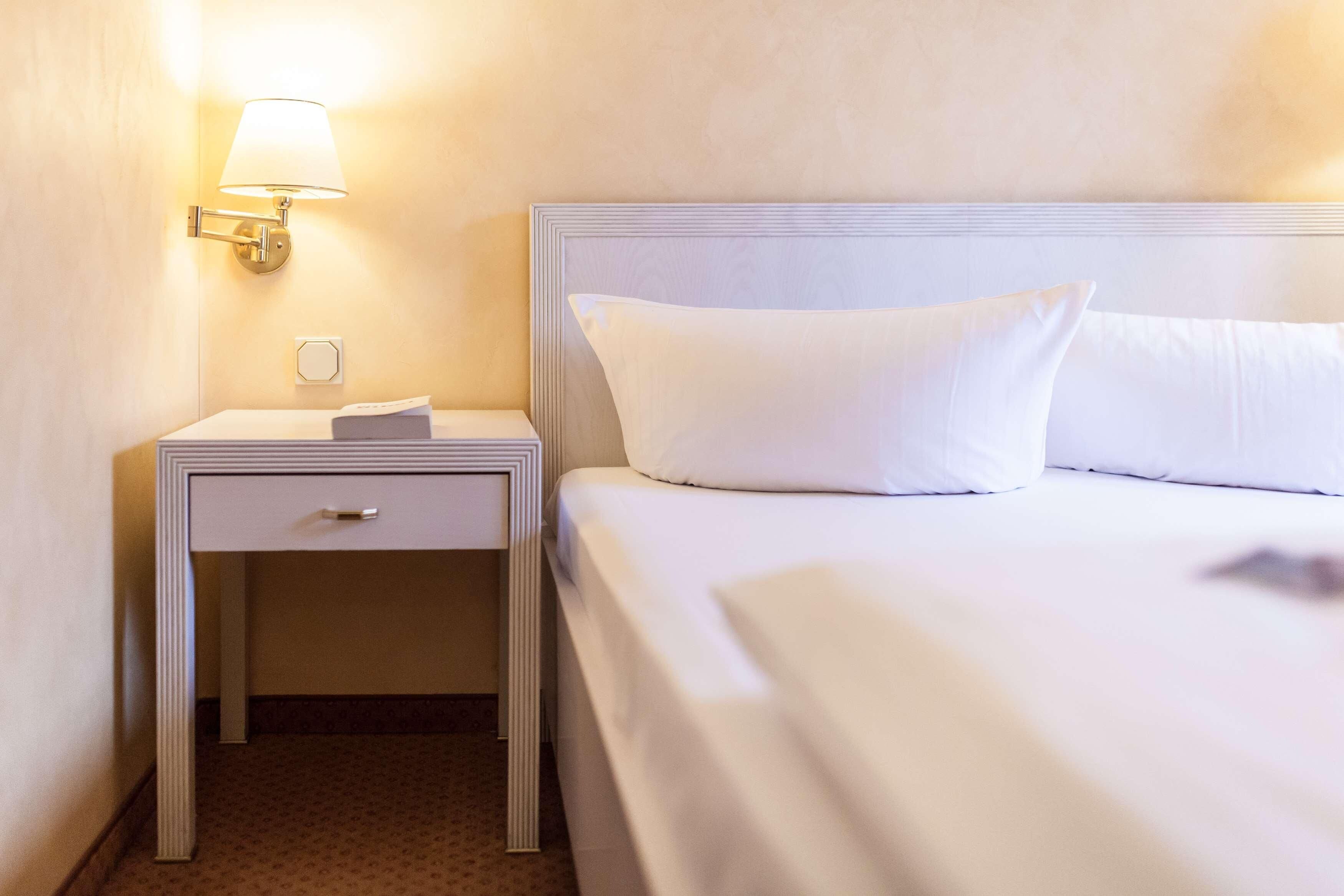 Ein Hotelzimmer mit einem ordentlich bezogenen Bett, einem Nachttisch mit Lampe und einer kleinen Schublade.