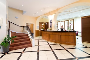 Uma área de recepção de hotel com um balcão de madeira, uma escadaria com guarda-corrimão preto e um piso de azulejos com padrão em preto e branco.