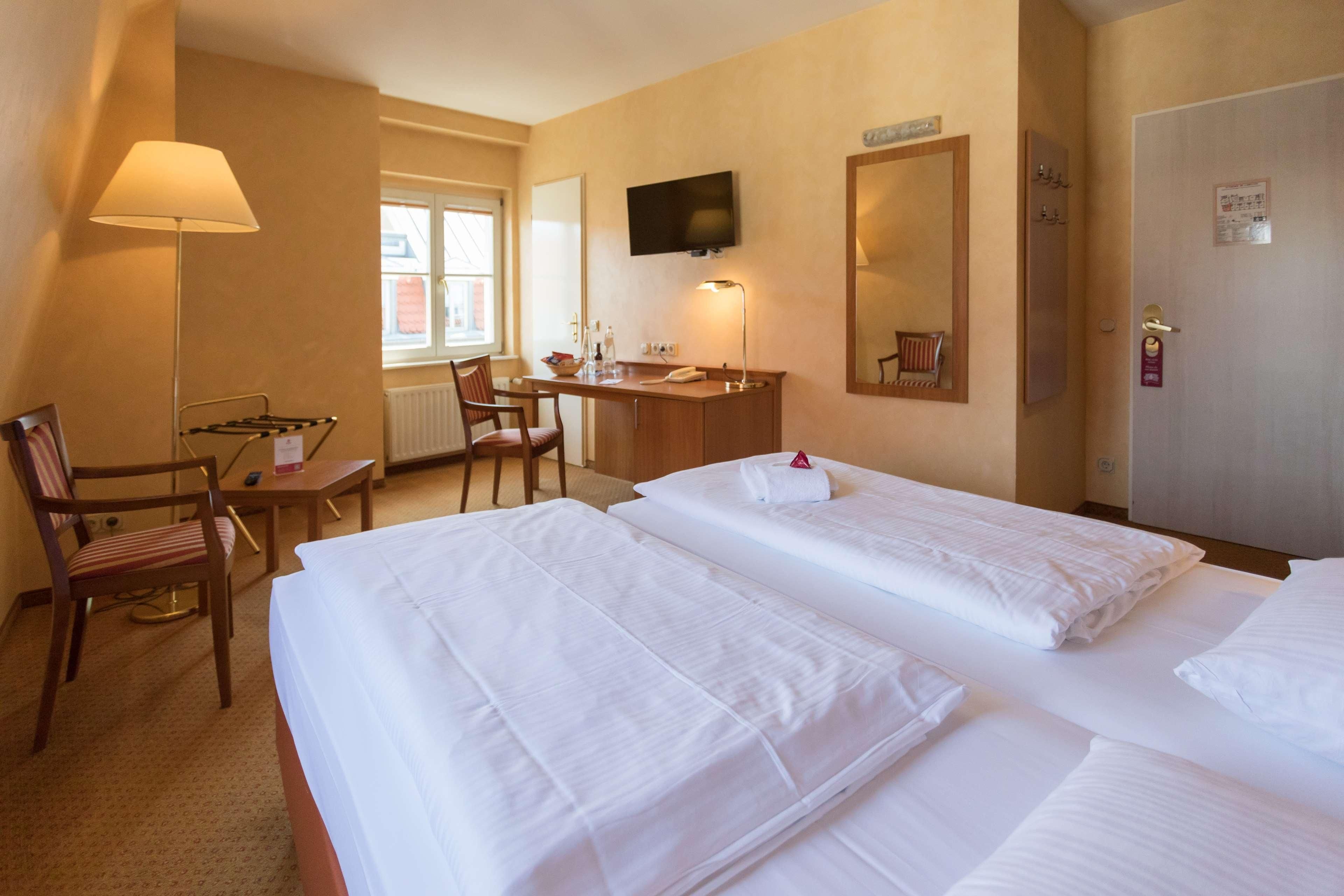 Ein Hotelzimmer mit zwei Betten, einem Schreibtisch, einem Stuhl, einem Fernseher und einem Fenster.