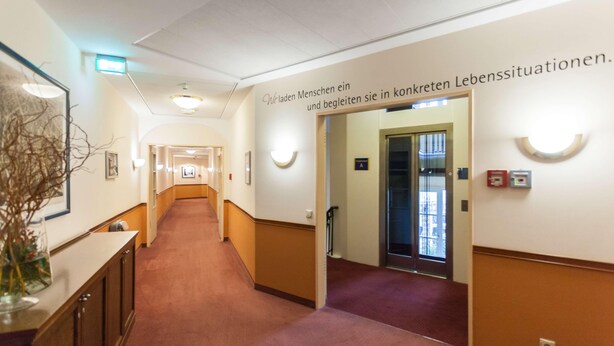 Un couloir doté d’un sol recouvert de moquette, un comptoir d’accueil orné d’un vase contenant des plantes séchées et un panneau indiquant les toilettes réservées aux femmes.
