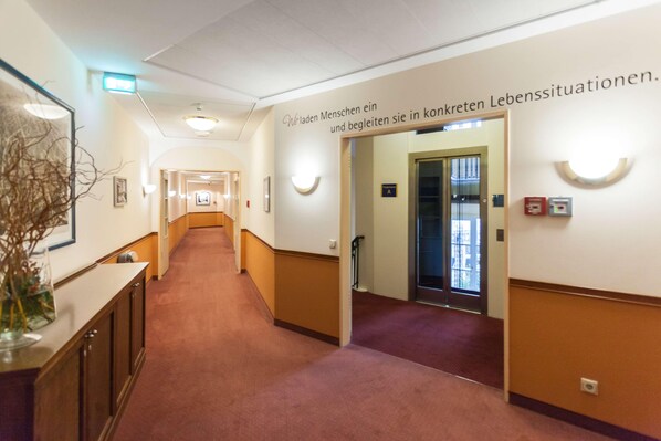 En gang med tæppebelagt gulv, en receptionsdisk med en vase tørrede planter, og et skilt der viser vejen til dametoilettet.