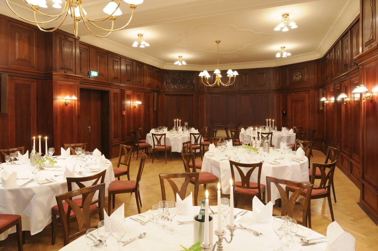 Ein Festsaal mit runden Tischen, Holzwandverkleidung, Kronleuchtern und Wandleuchten.