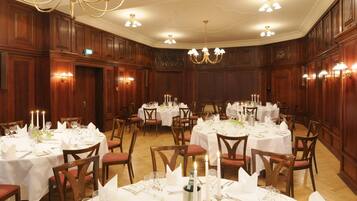 A banquet hall with round tables set for dining, wooden paneling, chandeliers, and wall sconces.