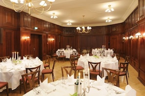 A banquet hall with round tables set for dining, wooden paneling, chandeliers, and wall sconces.