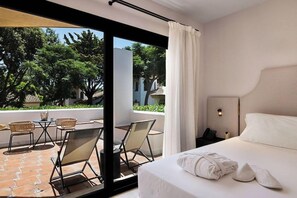 A hotel room with a bed, a nightstand, a phone, a window with a view of trees and a building, and a balcony with chairs and a table.