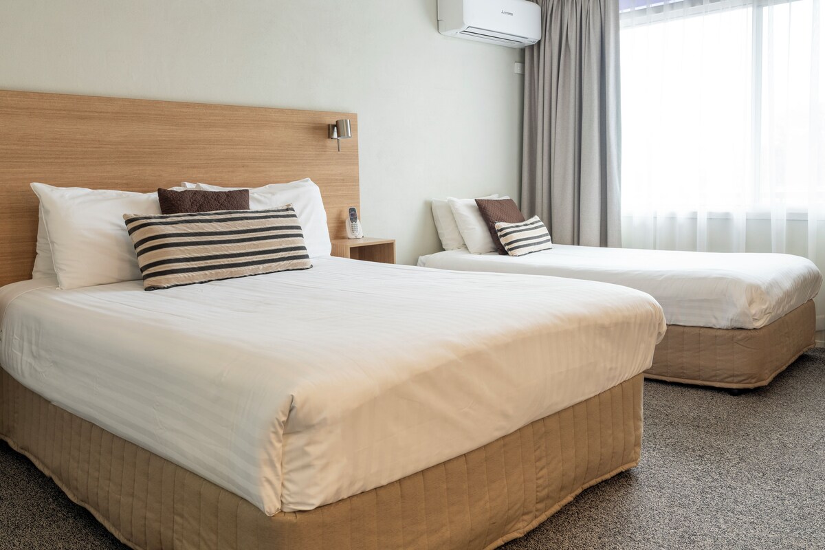 A hotel room with two beds, a wooden headboard, a bedside table with a lamp, and a window with curtains.