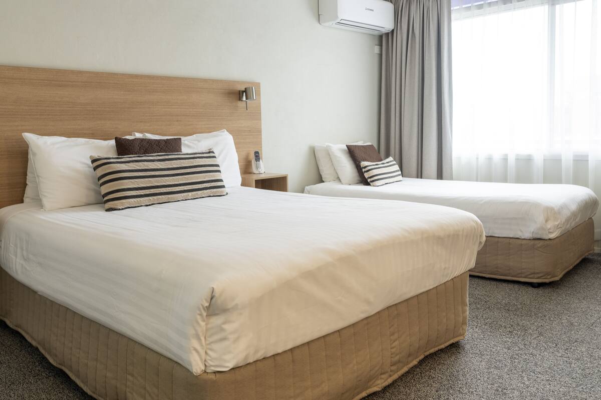 A hotel room with two beds, a wooden headboard, a bedside table with a lamp, and a window with curtains.
