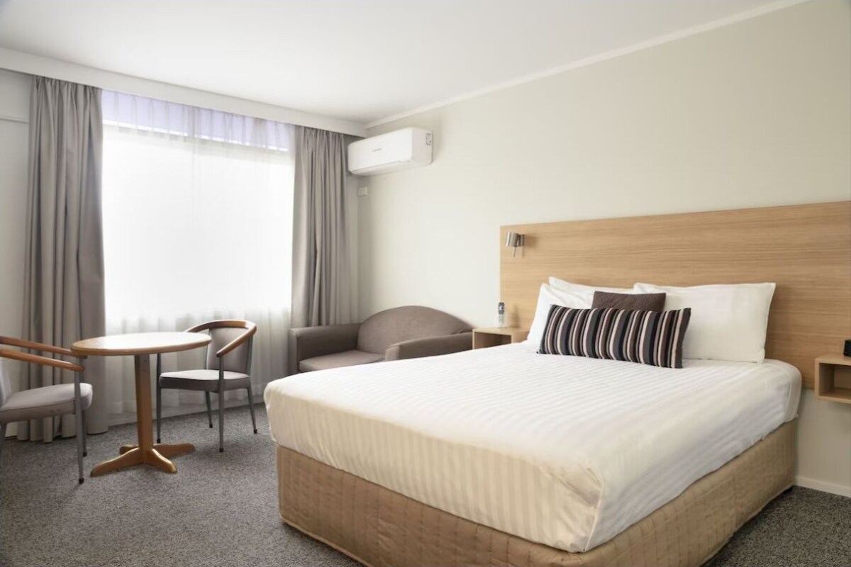 A hotel room with a large bed, a round table, two chairs, a desk, and a window with curtains.