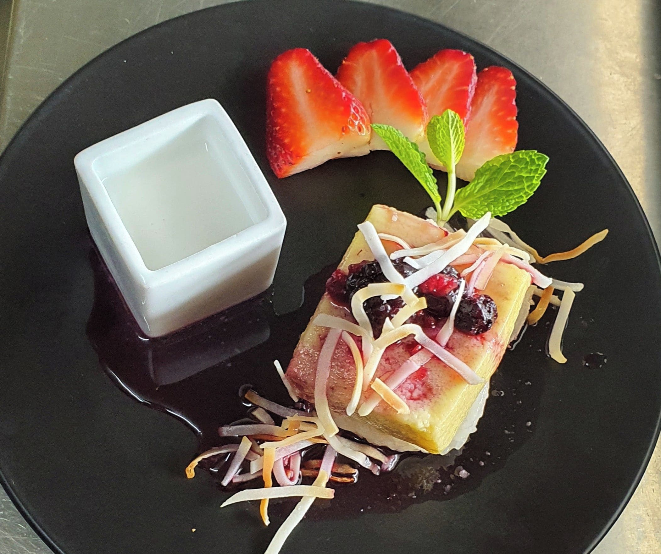 Un dessert à base de riz gluant, de crème pâtissière thaïlandaise, de fraises et de feuilles de menthe, présenté sur une assiette noire.