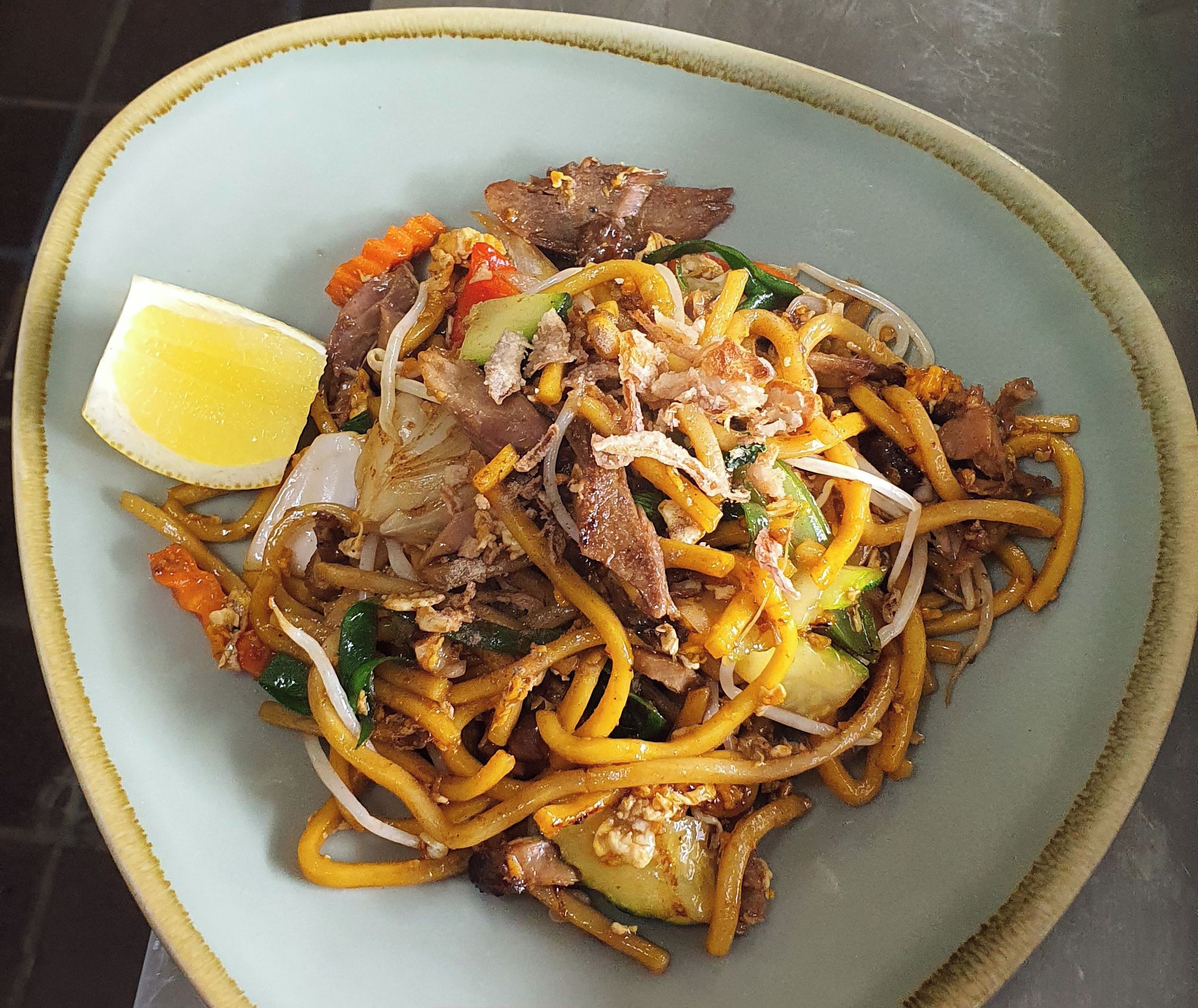 A plate of Hokkien Duck noodles with vegetables and a lemon wedge.