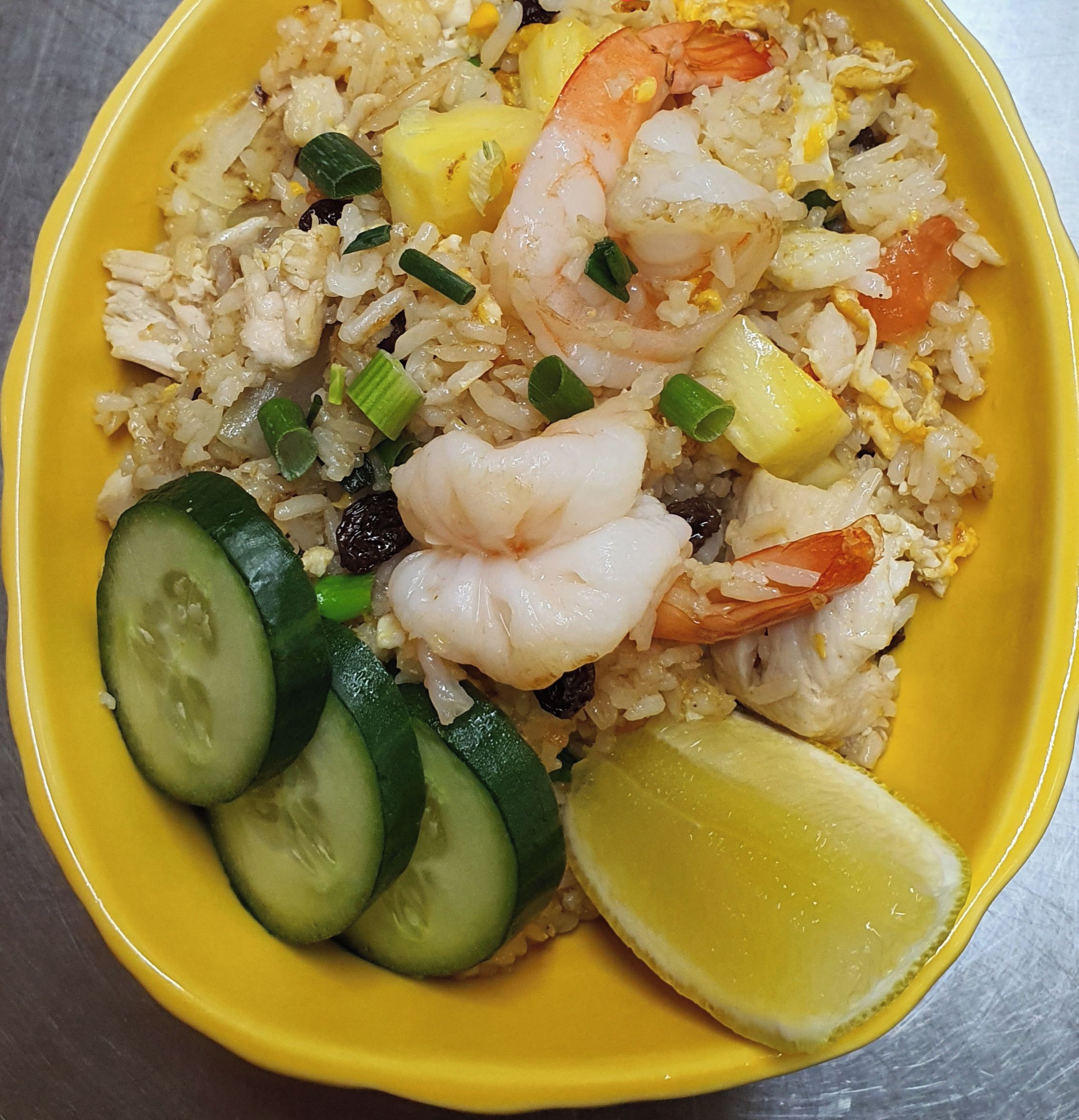 A yellow bowl of pineapple fried rice with chicken and prawns, garnished with cucumber slices and a lime wedge.