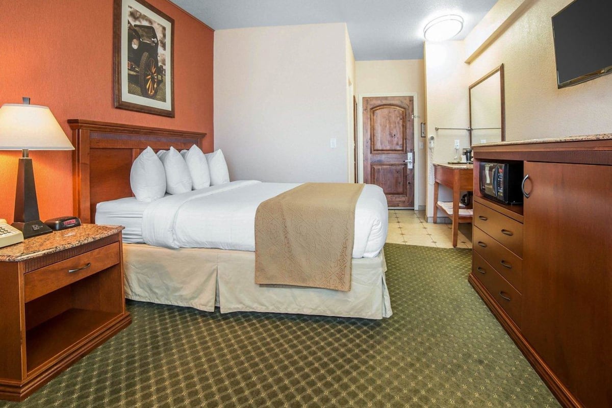 A hotel room with a bed, a desk with a lamp, a dresser, a microwave, and a door leading to another room.