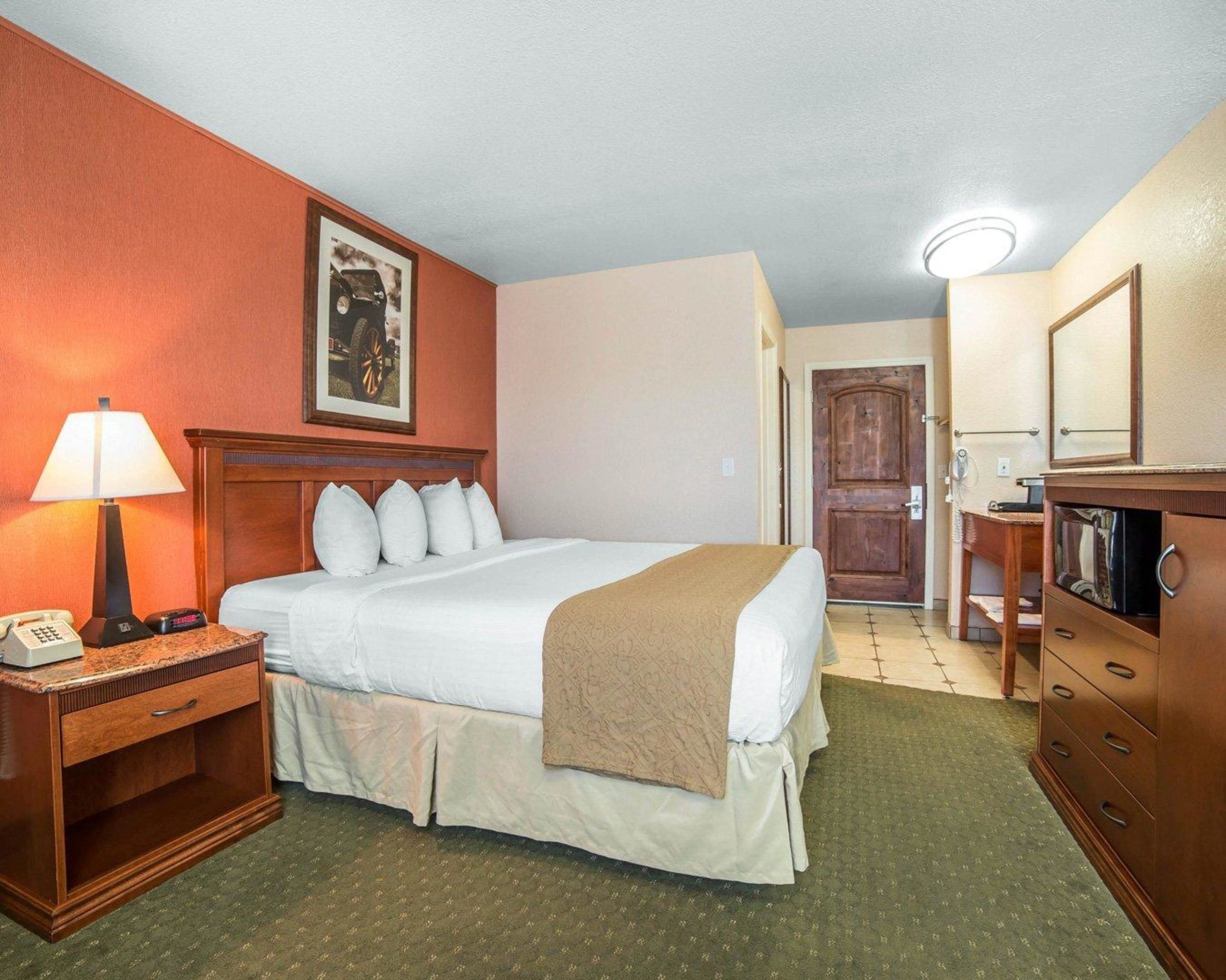 A hotel room with a bed, bedside table with a lamp, a dresser, a mirror, and a door leading to another room.