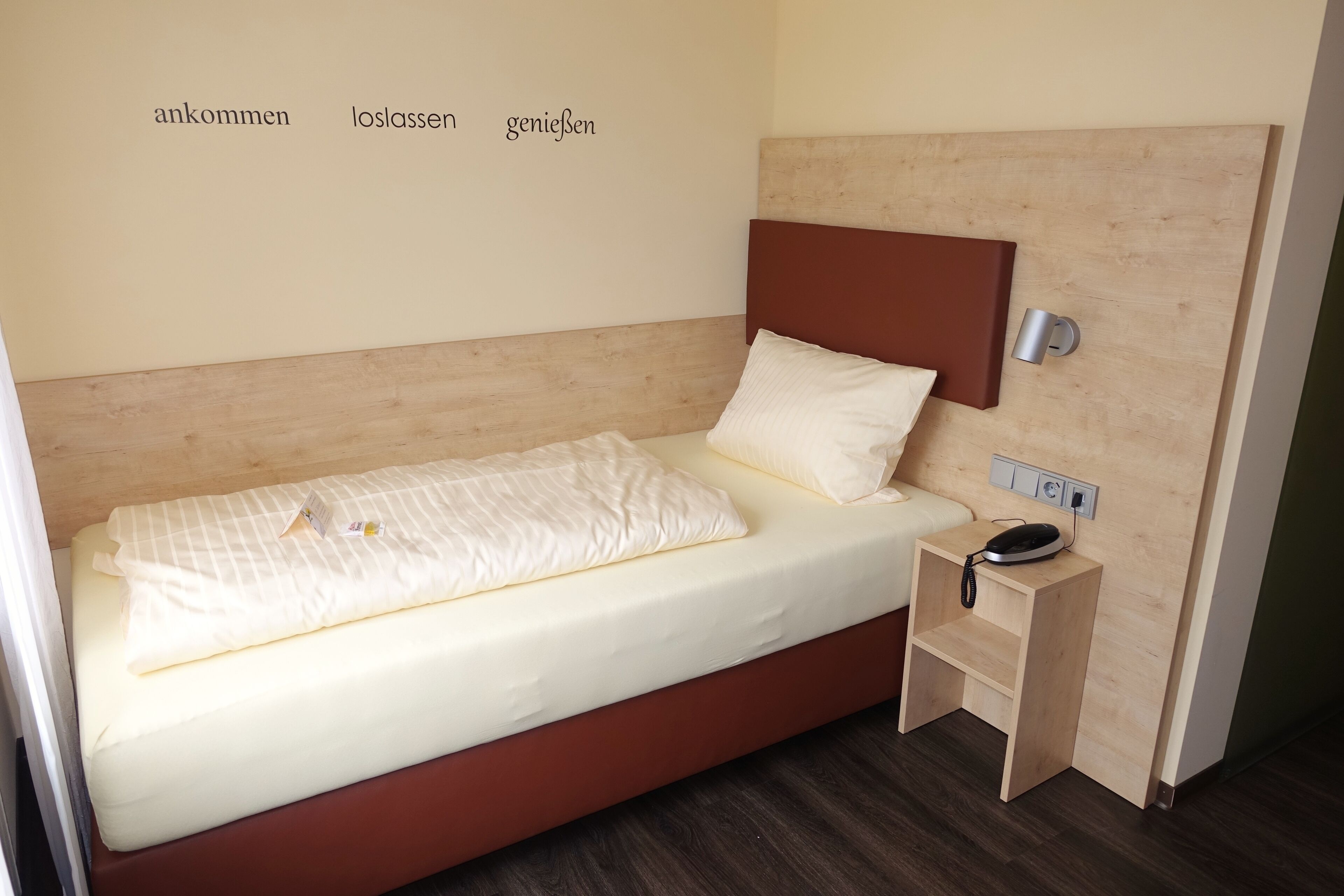 A single bed with a wooden headboard, a bedside table with a lamp, and a wall with text.