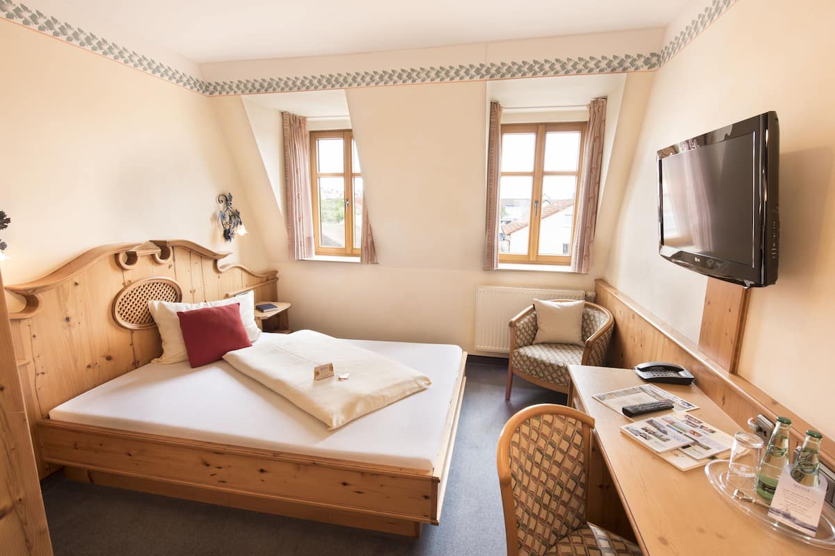 Une chambre à coucher avec un lit en bois, un bureau avec une chaise, une télévision et deux fenêtres avec des rideaux.