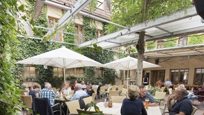 Un café en plein air, avec des tables et des chaises, entouré de verdure et bordé d’un bâtiment dont le toit est recouvert de tuiles.