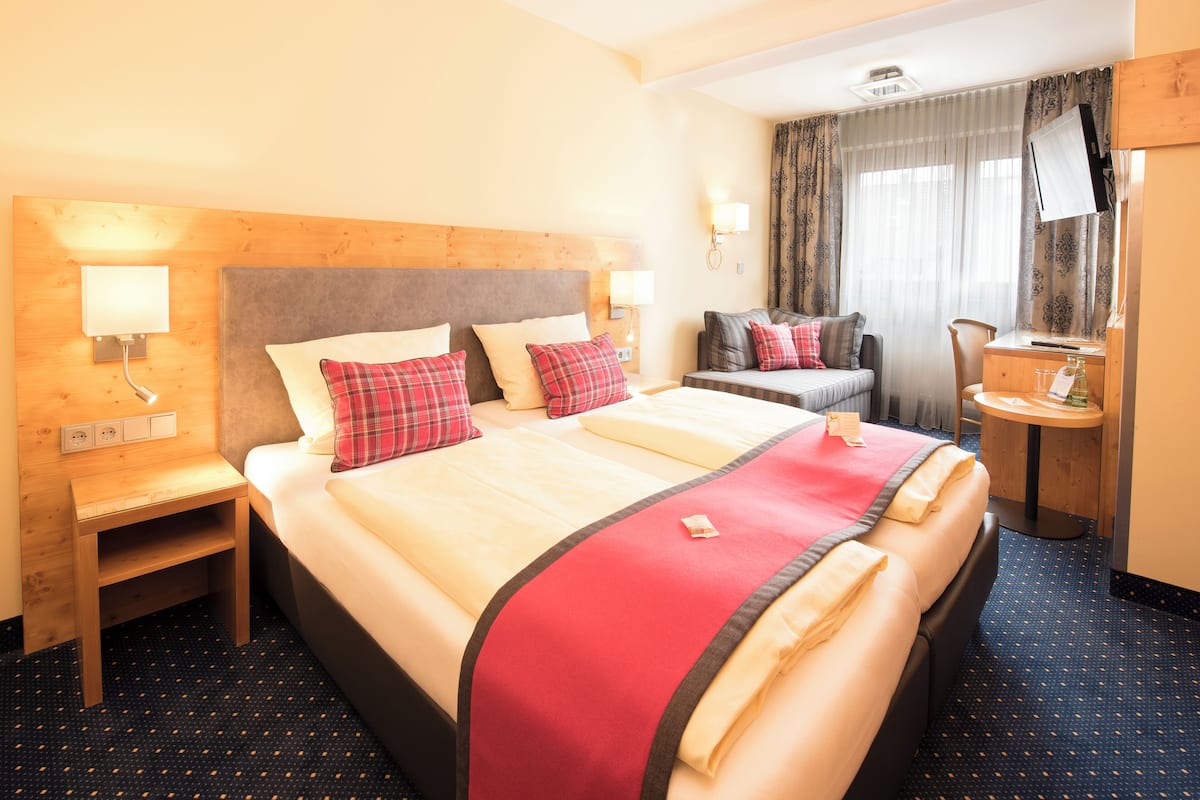 Une chambre d’hôtel avec un grand lit, une table de chevet, un bureau avec une chaise, un miroir et une fenêtre avec des rideaux.