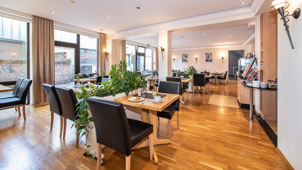 A modern dining area with wooden tables and black chairs, a coffee station, and large windows with curtains. - UNO Hotel Posthof Saarlouis by ANS (Saarlouis)
