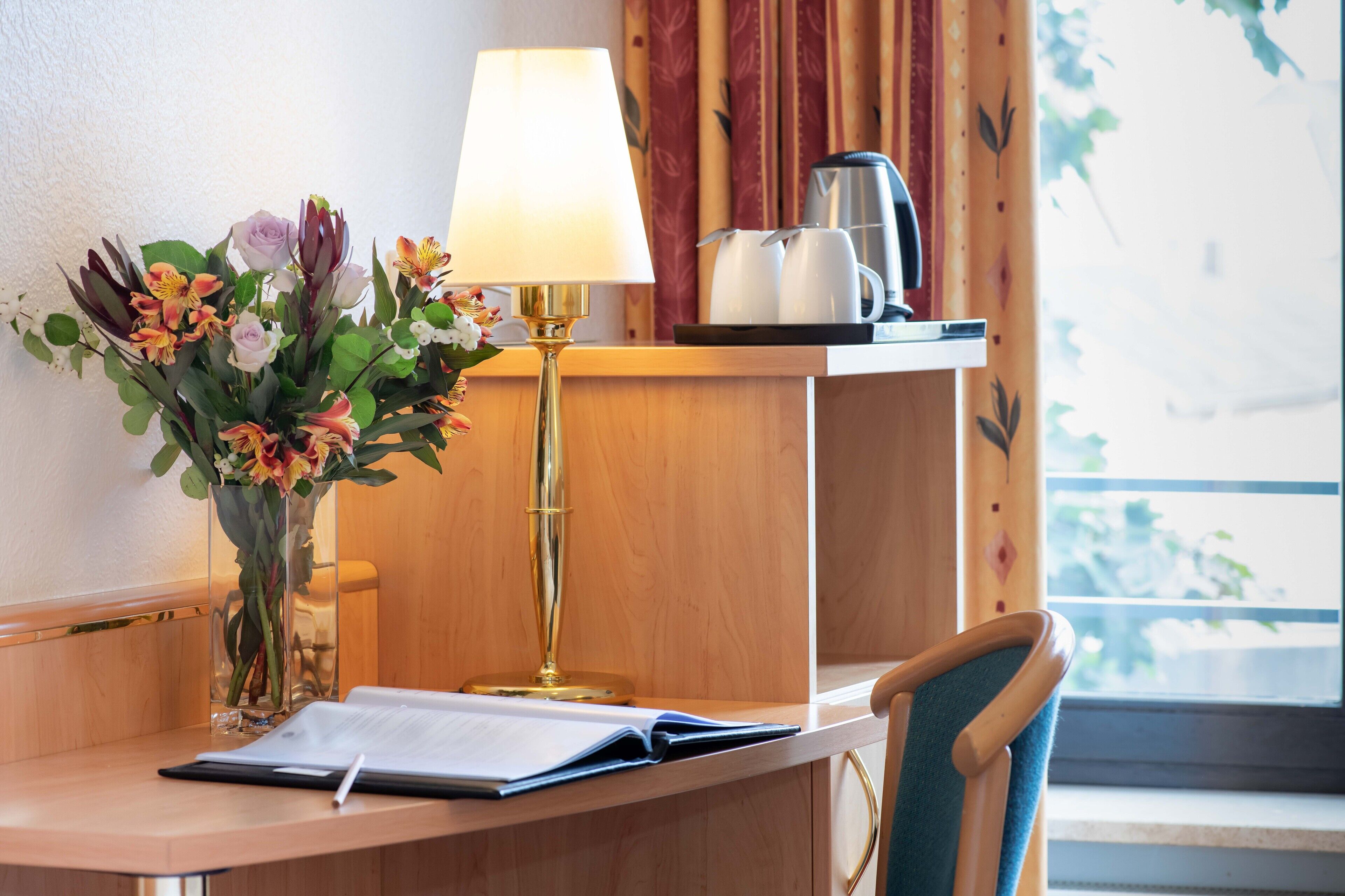 Un bureau dans une chambre d’hôtel, avec un vase de fleurs, une lampe, une bouilloire et une chaise.