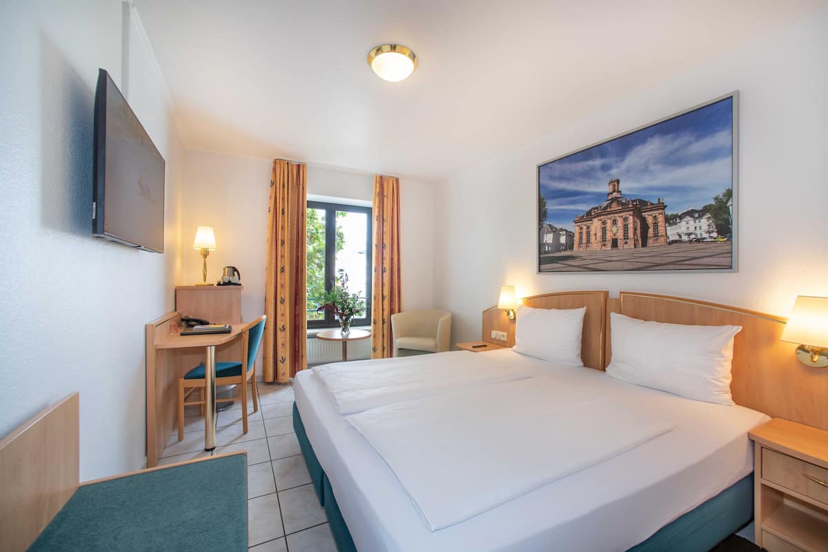Une chambre d’hôtel équipée d’un lit, d’un bureau, d’une chaise, d’une lampe et d’un tableau représentant un bâtiment.