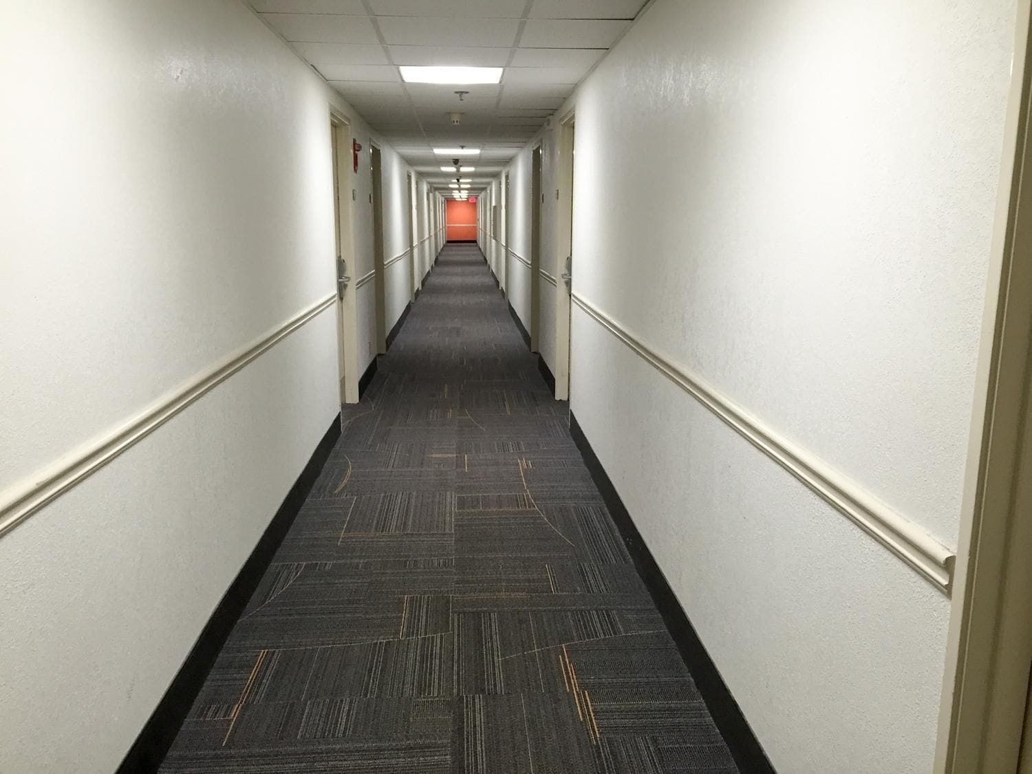 Un long couloir étroit, typique d’un hôtel, avec un sol recouvert de moquette et des murs blancs.