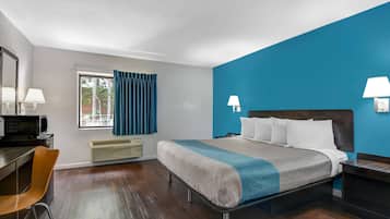 A hotel room with a bed, a desk, a chair, a window with curtains, and a view of trees.